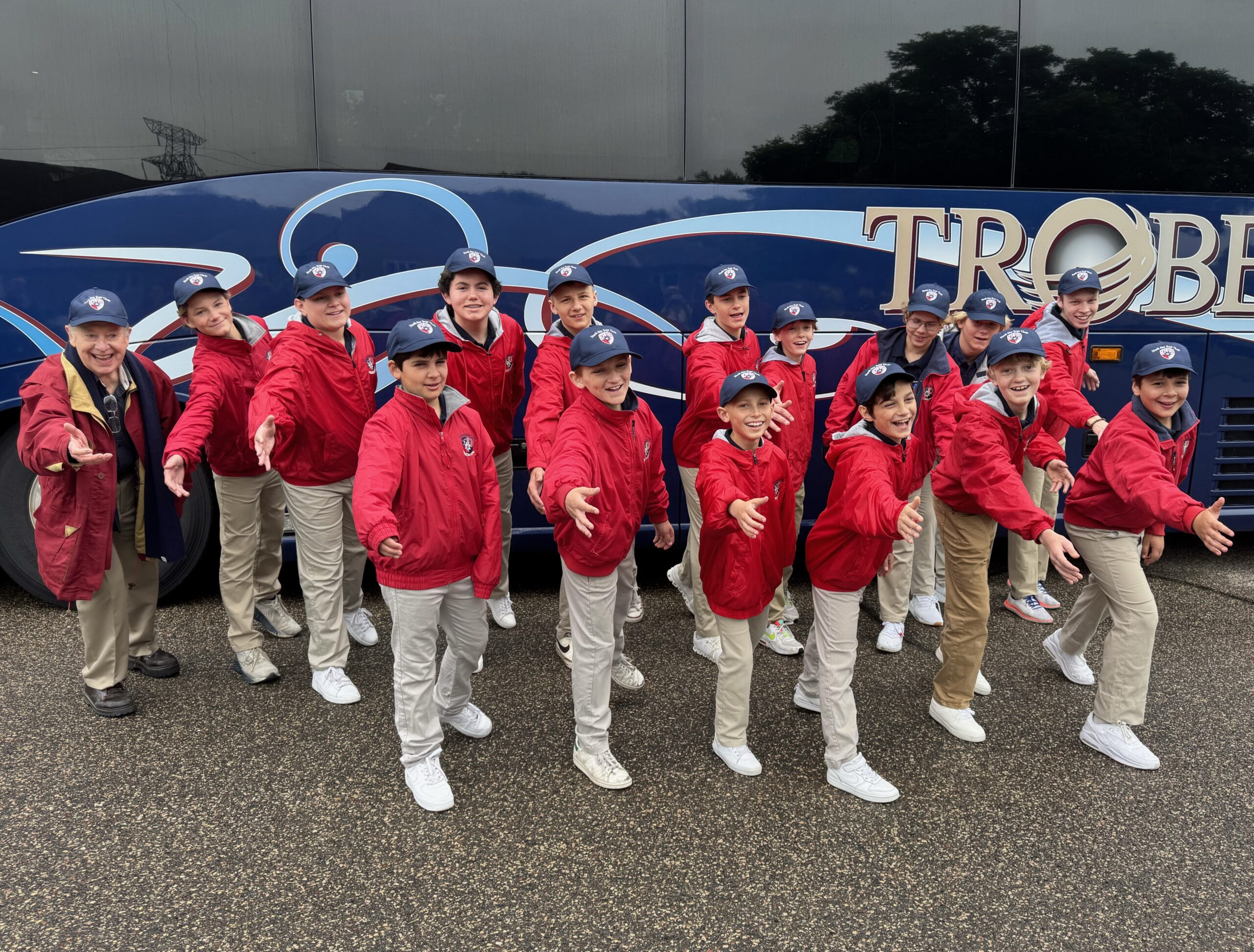 North Star Boys’ Choir members and leaders in red jackets and navy caps posing in front of a blue charter bus in a parking lot